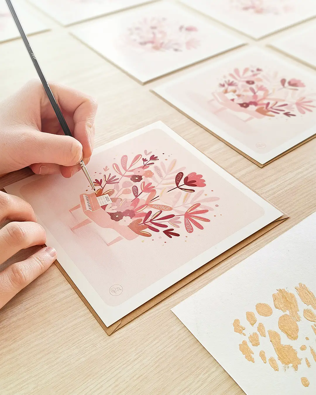Illustratrice en train d'appliquer des pointes de dorure sur des cartes illustrées, représentant un piano à queue avec des feuilles, fleurs et messages positifs.