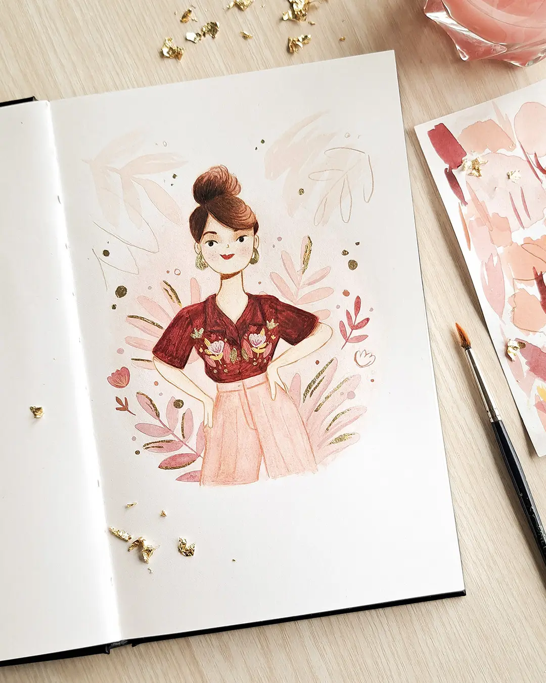 Page de carnet de croquis avec l'illustration d'une femme avec un chemisier imprimé, entourée de feuilles tropicales et de petits éclats de feuilles d'or.