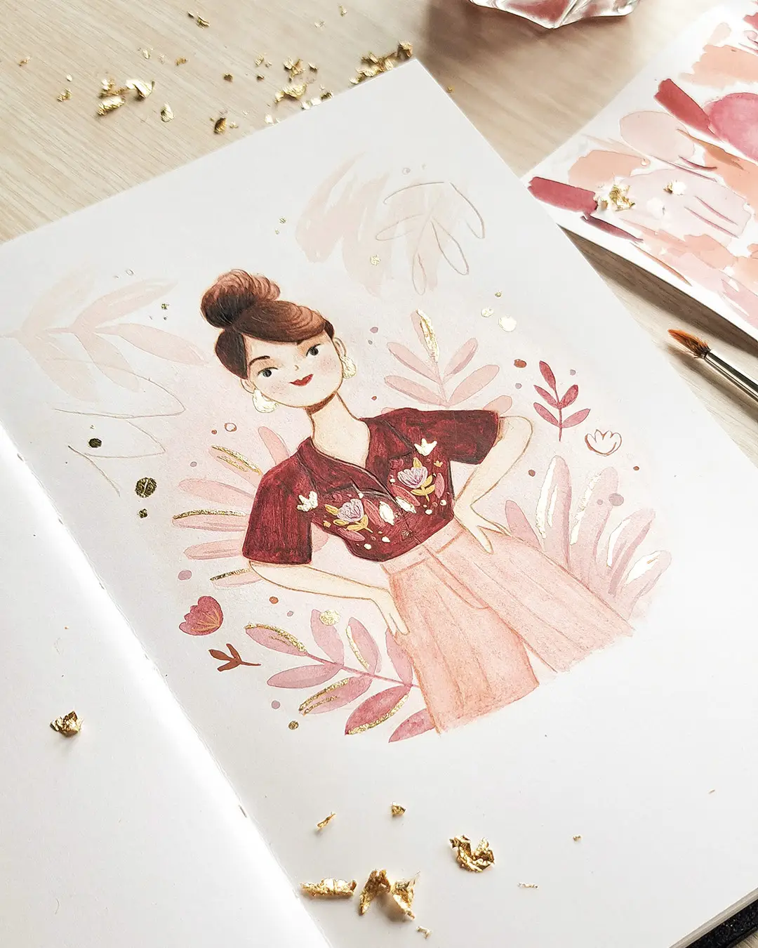 Page de carnet de croquis avec l'illustration d'une femme avec un chemisier imprimé, entourée de feuilles tropicales et de petits éclats de feuilles d'or.